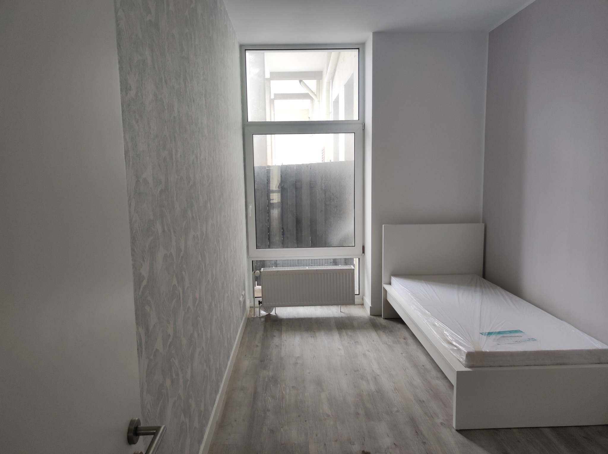 Pomalowane na szaro ściany pokoju z wzorzystą tapetą, białe łóżko z materacem w folii, grzejnik pod oknem i panele na podłodze.