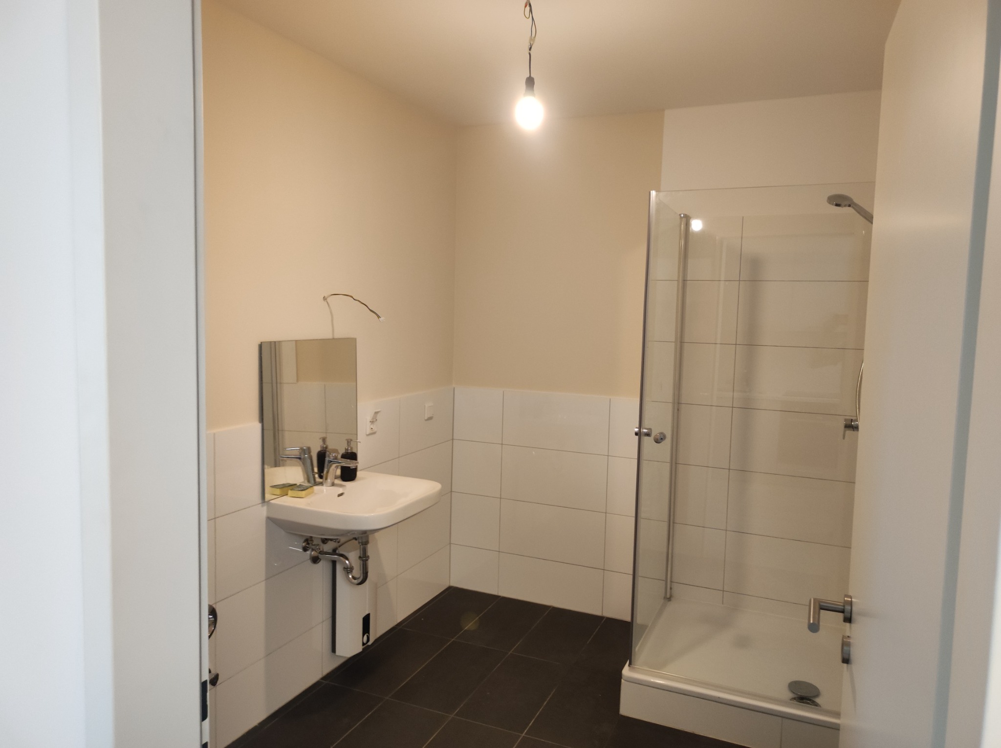 Nowoczesna łazienka z białymi płytkami, ciemną podłogą, kabiną prysznicową ze szklanymi drzwiami i umywalką z lustrem, oświetlona pojedynczą żarówką wiszącą na kablu.