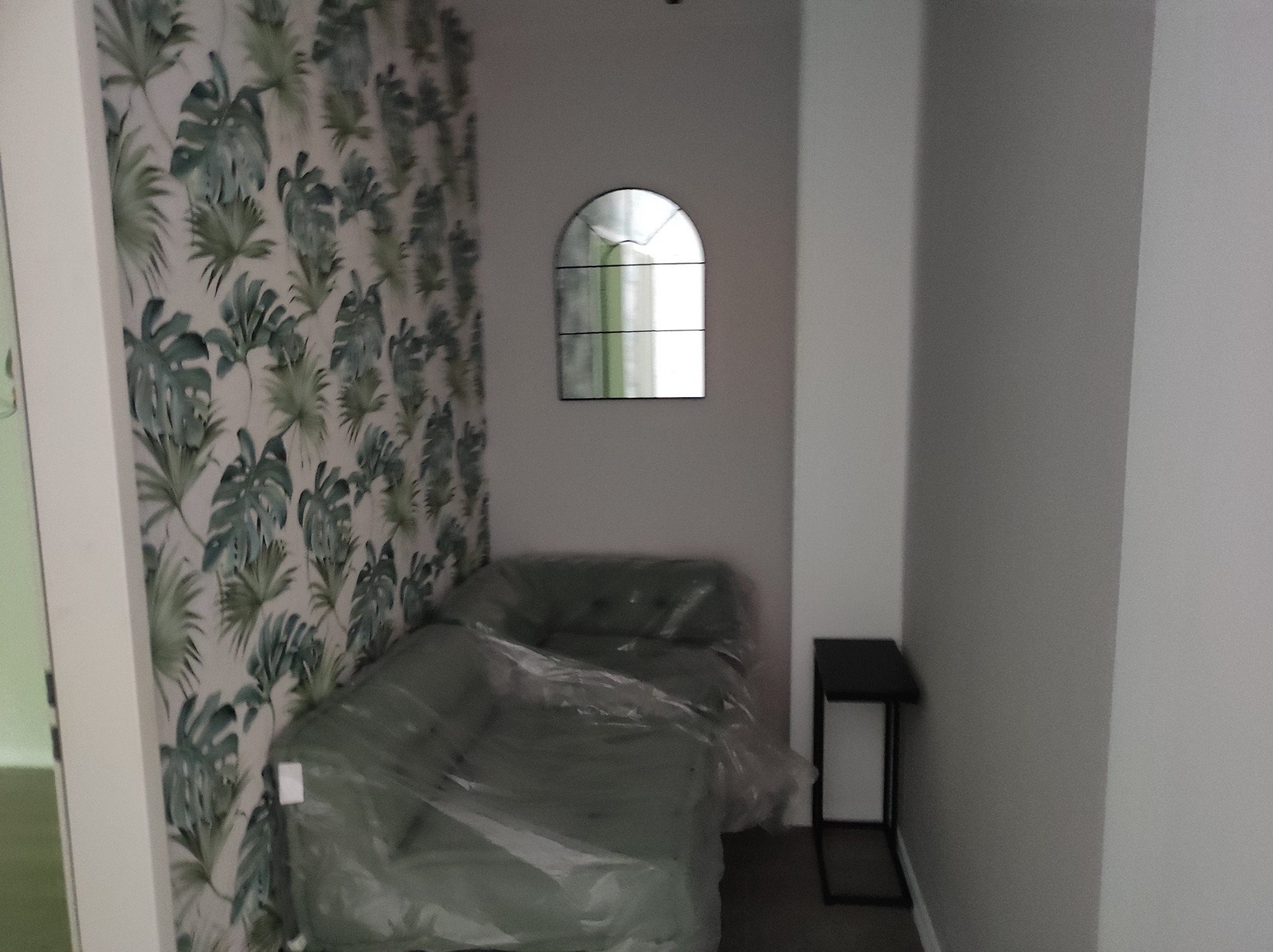 Pokój po odświeżeniu: ściana z tapetą w liście, lustro w kształcie okna, sofa zabezpieczona folią ochronną i mały stolik pomocniczy.