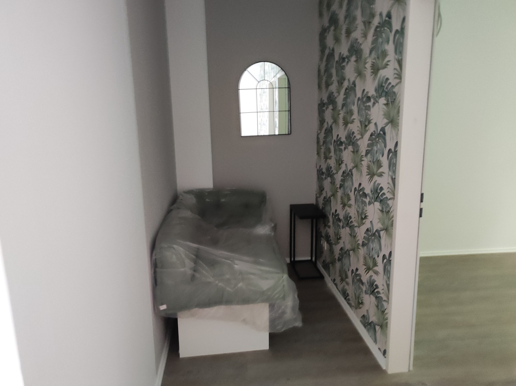 Pokój po remoncie: ściana z tapetą w liście, lustro w kształcie okna, mebel przykryty folią ochronną, mały stolik.