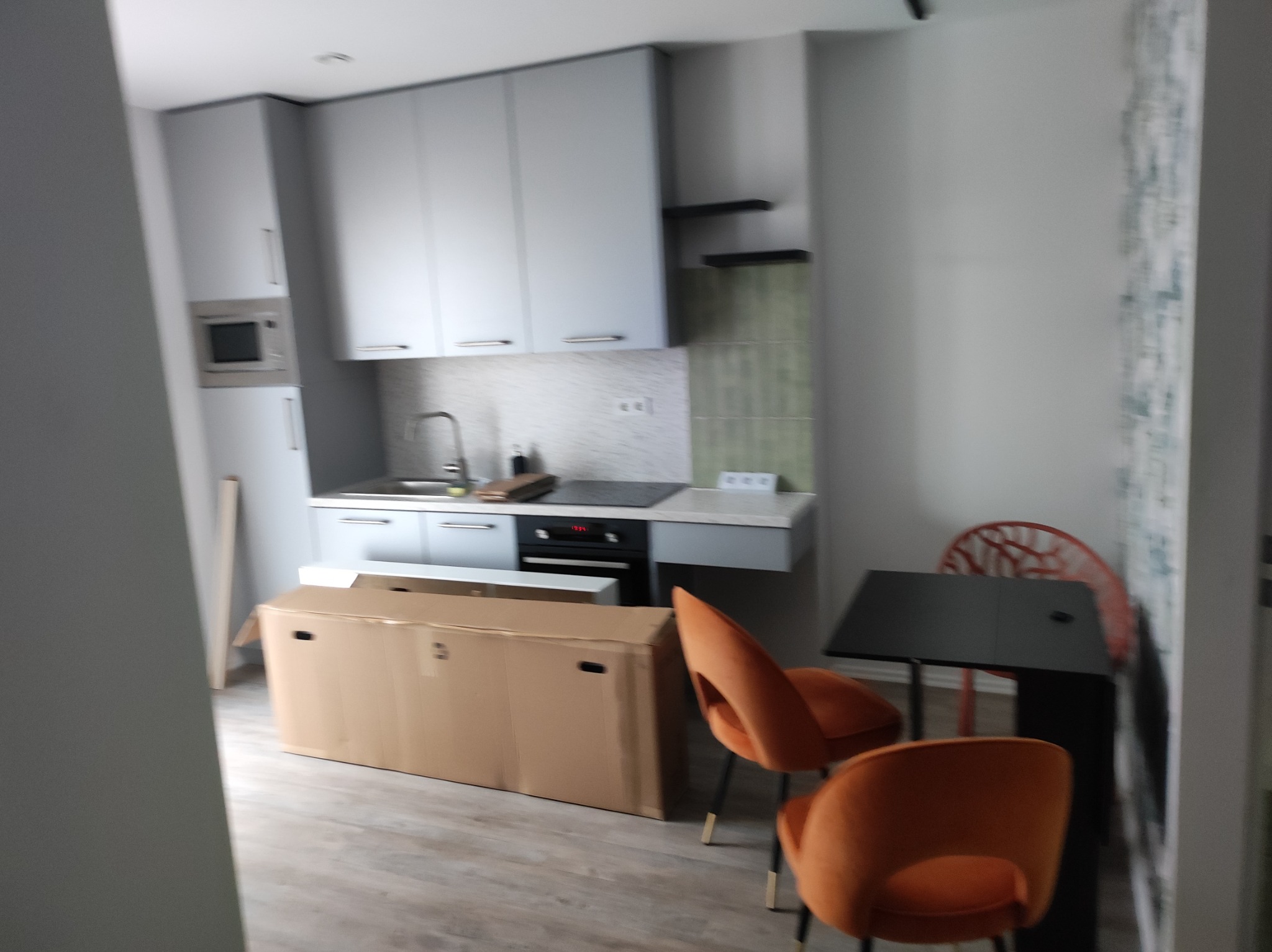 Wnętrze mieszkania w trakcie urządzania: kartonowe opakowanie mebli stoi na podłodze obok pomarańczowych krzeseł i stołu, widoczny fragment zabudowy kuchennej w szarych odcieniach.