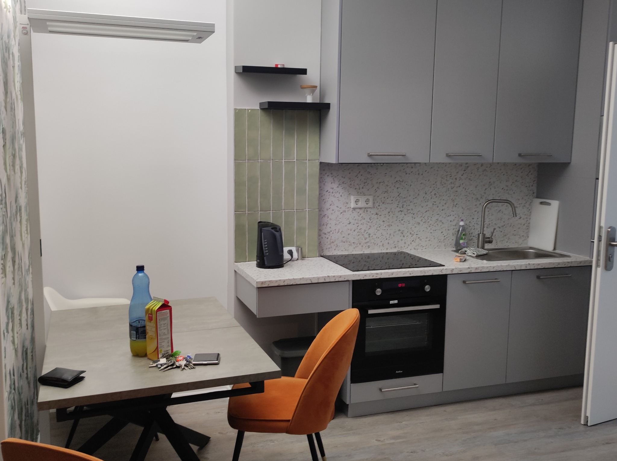 Nowoczesna kuchnia w odcieniach szarości z pomarańczowym krzesłem, na stole butelka wody, sok, portfel, klucze i telefon, płytki na ścianie w odcieniu pistacjowym.