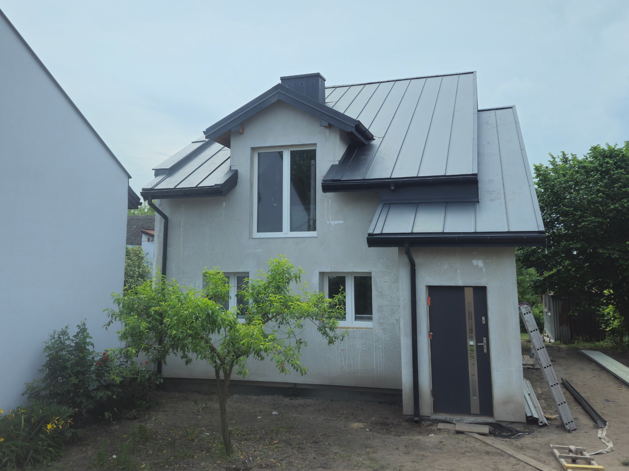 Dom jednorodzinny z nowym, metalowym dachem w kolorze grafitowym, widoczne obróbki blacharskie i drabina oparta o ścianę budynku.