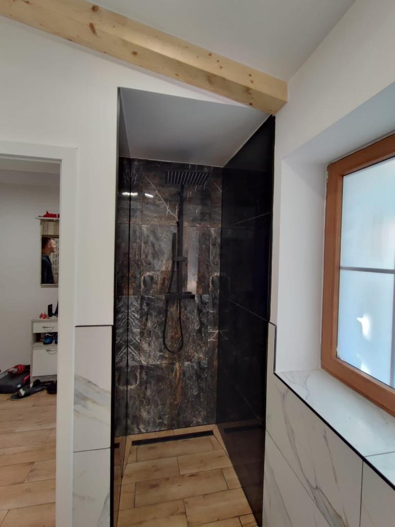 Nowoczesna kabina prysznicowa z czarnymi płytkami imitującymi marmur, drewnianą podłogą i widoczną belką stropową w łazience z oknem.
