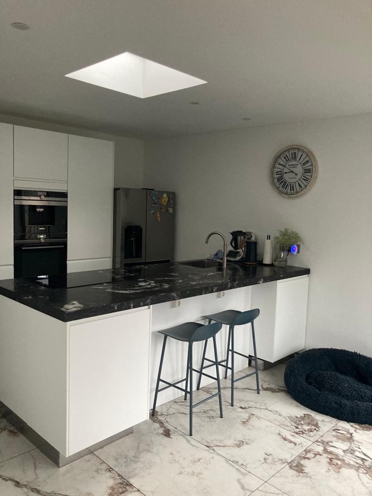 Nowoczesna kuchnia z wyspą o czarnym, nakrapianym blacie, białymi szafkami i marmurową podłogą, oświetlona przez świetlik dachowy. Dwa niebieskie hokery stoją przy wyspie, a obok leży ciemne...