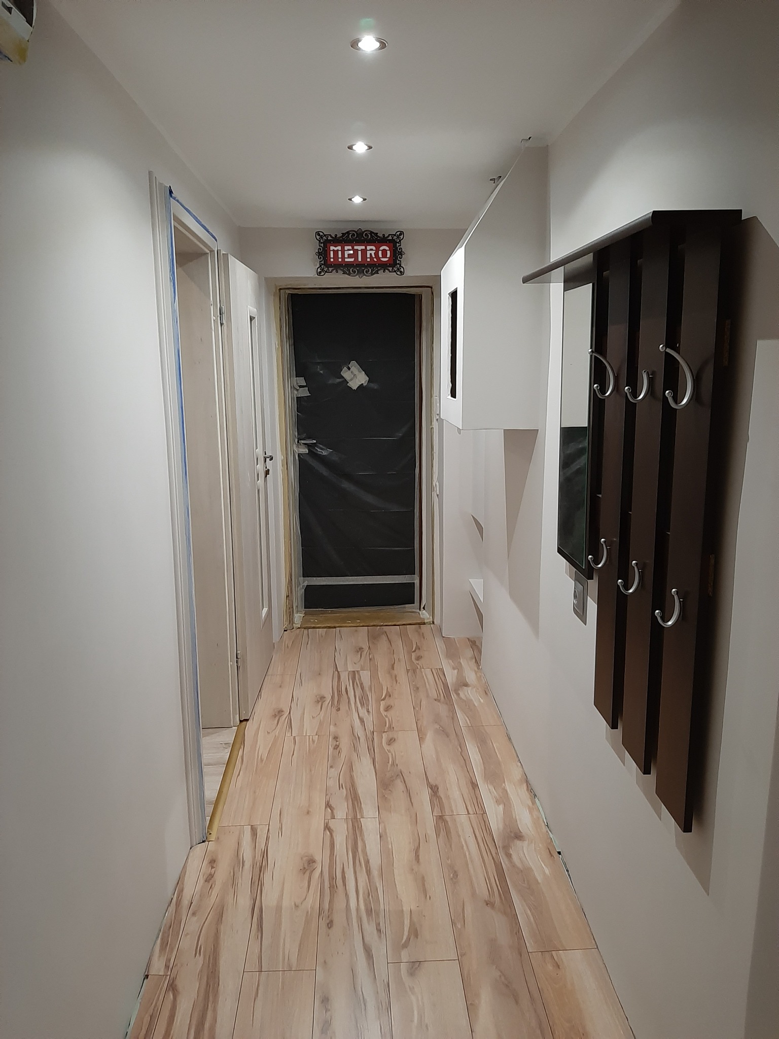 Korytarz w trakcie odświeżania: widoczne zabezpieczenia folią drzwi, świeżo pomalowane ściany, drewnopodobna podłoga i wieszak z lustrem.