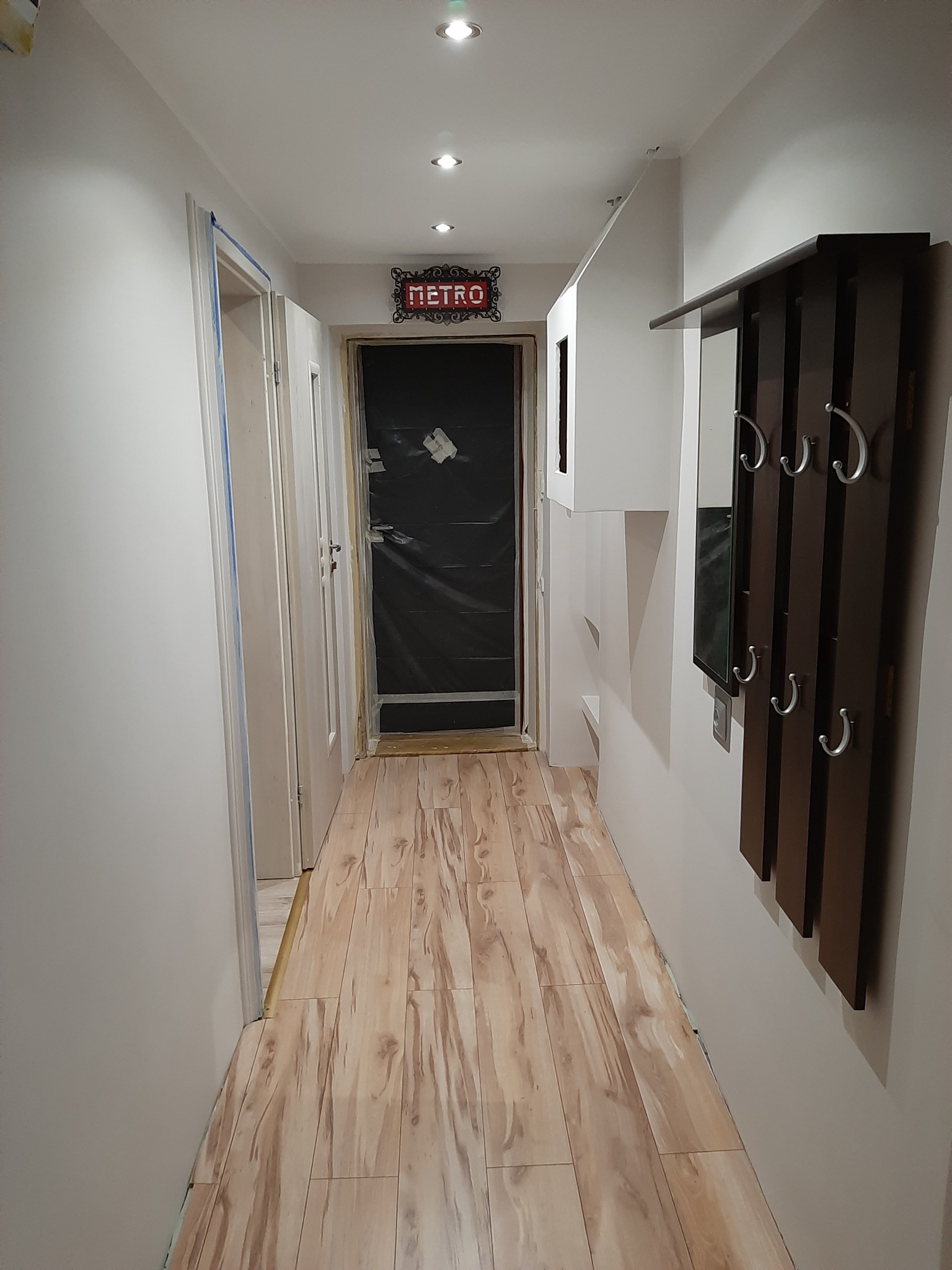 Korytarz po remoncie z nową podłogą imitującą drewno, pomalowanymi na biało ścianami, oraz wieszakiem z lustrem. Drzwi zabezpieczone folią malarską.
