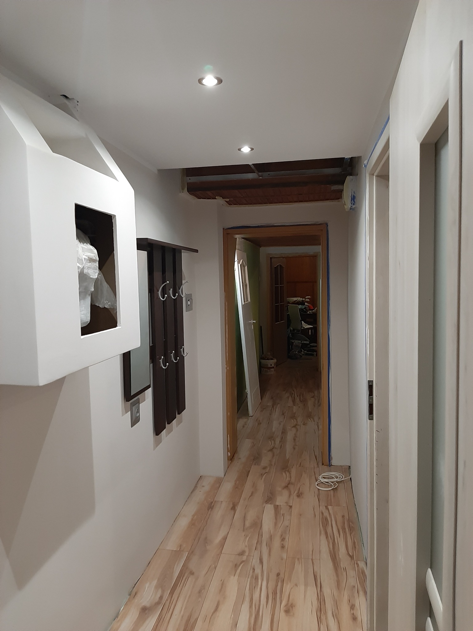 Korytarz w trakcie prac wykończeniowych: świeżo pomalowane ściany na biało, widoczne drewnopodobne panele na podłodze, niezamaskowane fragmenty sufitu i wystające kable. Na ścianie wisi biała...