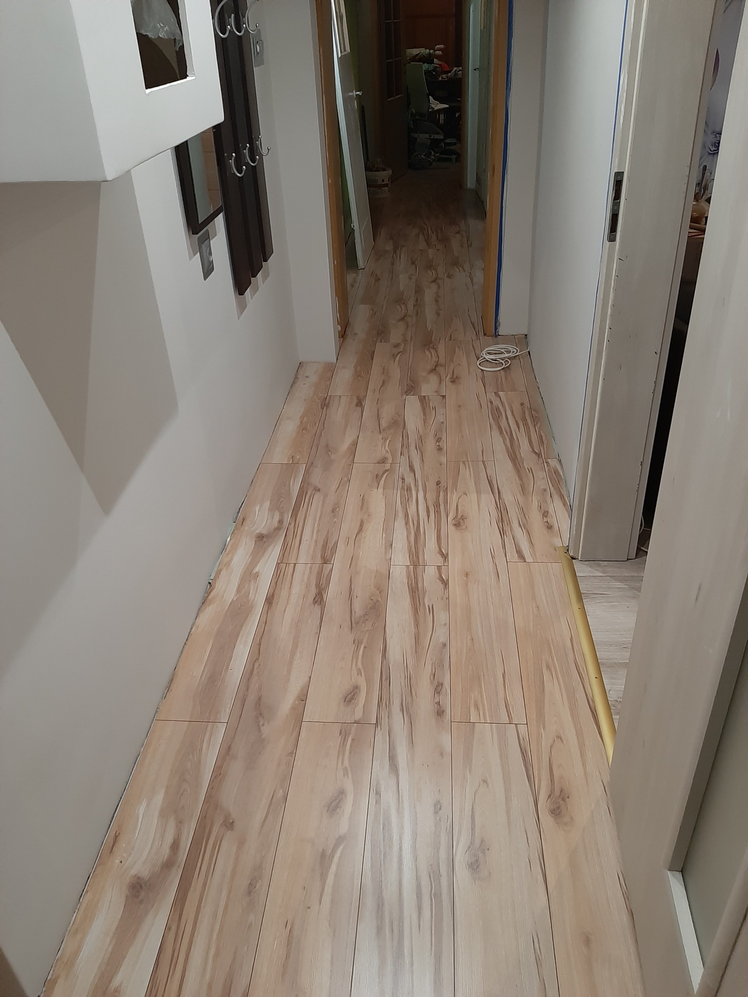 Ułożona podłoga z paneli imitujących jasne drewno w wąskim przedpokoju, widoczne białe ściany i drzwi oraz elementy wyposażenia wnętrza.