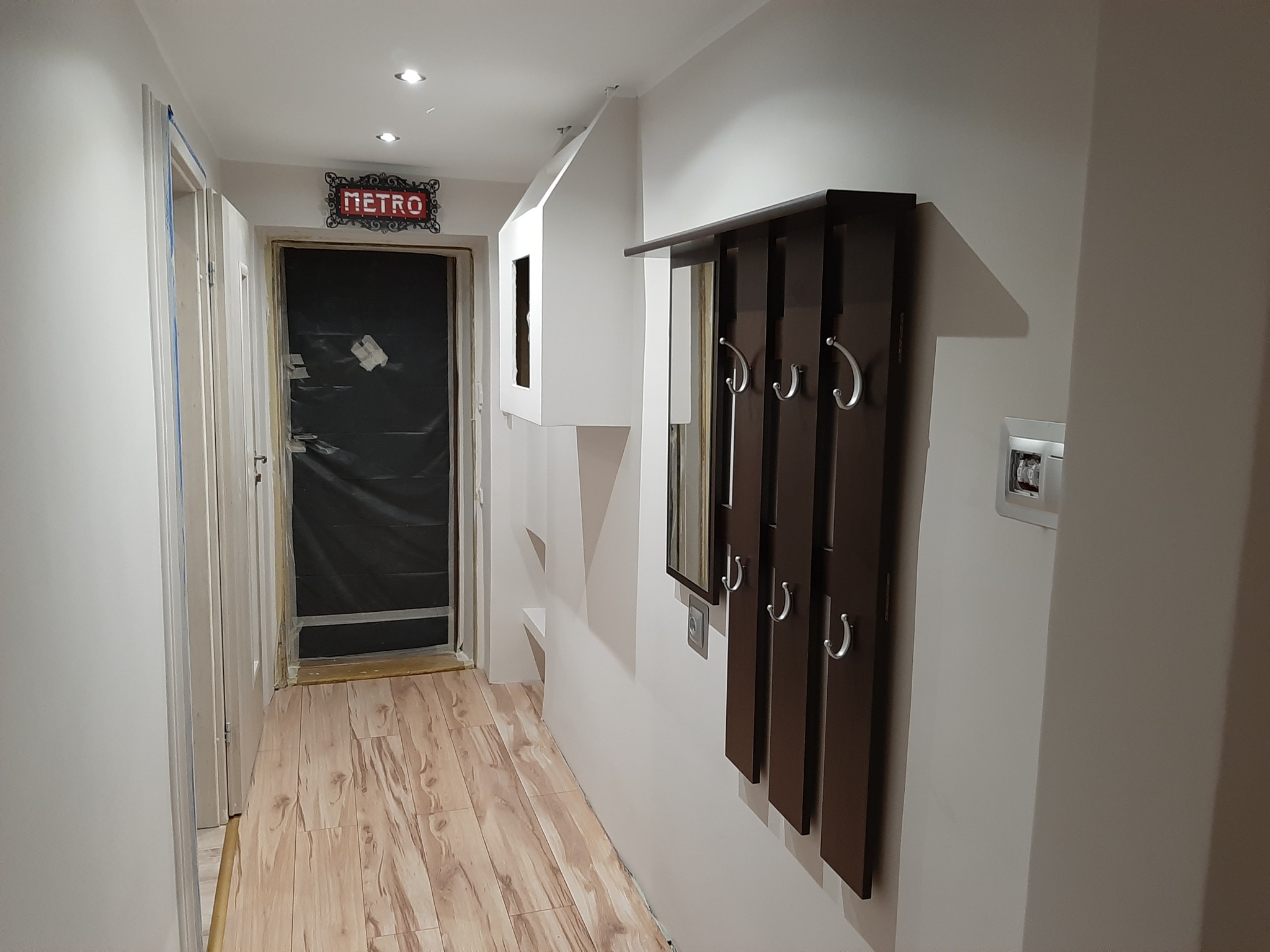 Przedpokój w trakcie remontu z zasłoniętymi folią drzwiami, drewnianą podłogą, wieszakiem z lustrem i dekoracją ścienną w stylu retro.