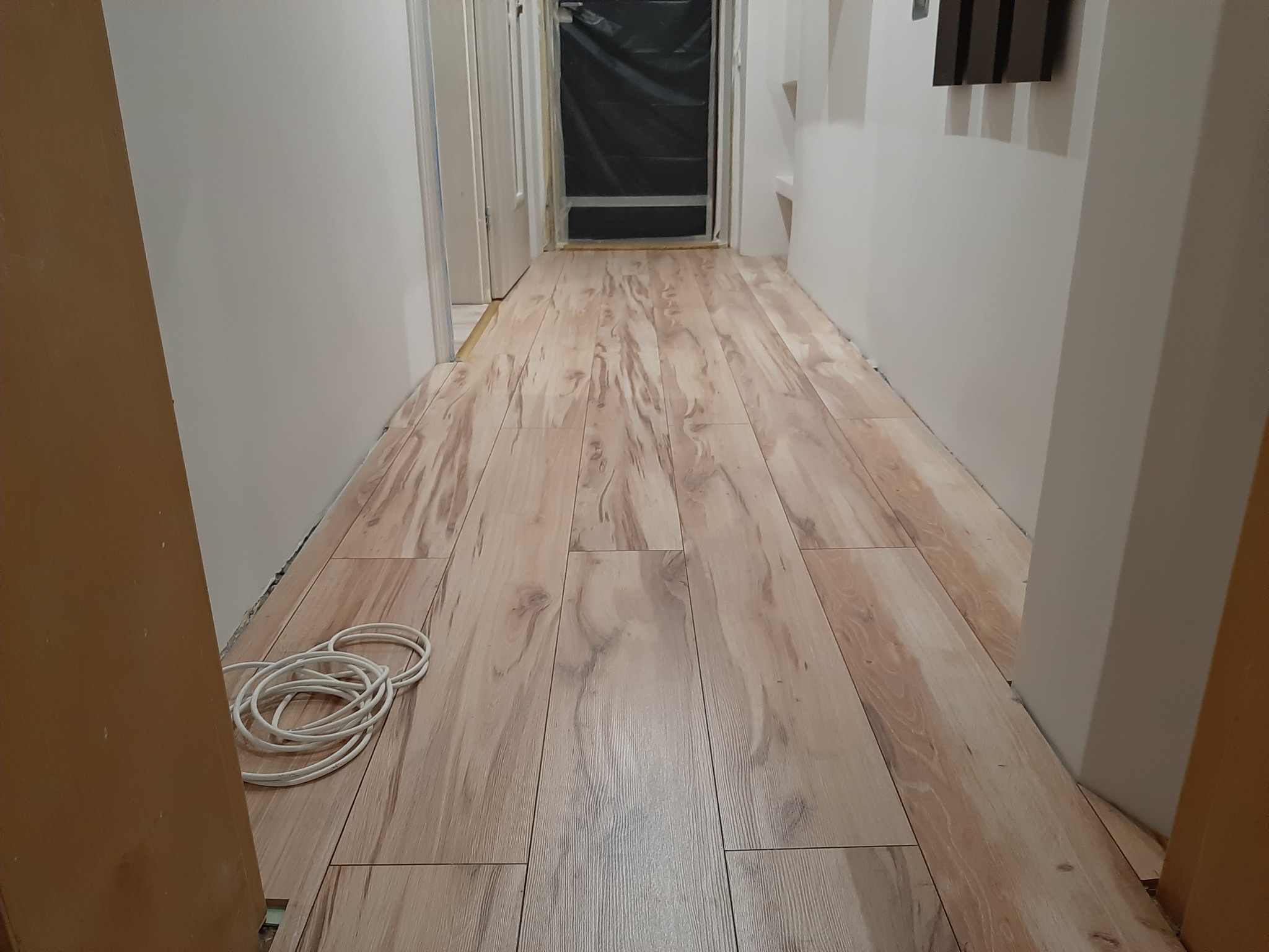 Korytarz w trakcie układania paneli podłogowych imitujących drewno, zabezpieczone drzwi folią ochronną, widoczne kable elektryczne przy ścianie.