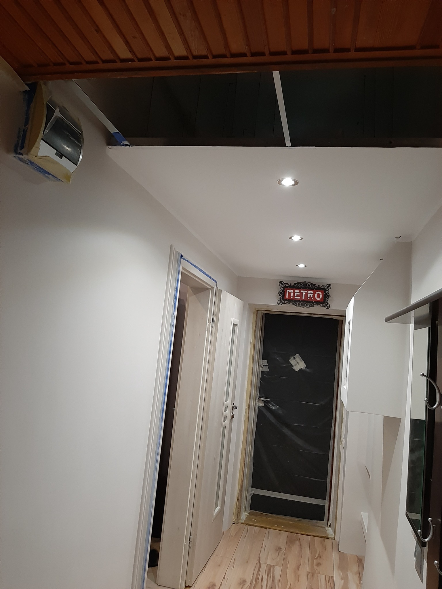 Korytarz w trakcie prac wykończeniowych: ściany pomalowane na biało, drzwi i klimatyzator zabezpieczone taśmą malarską, widoczne oświetlenie sufitowe i drewniana podłoga.