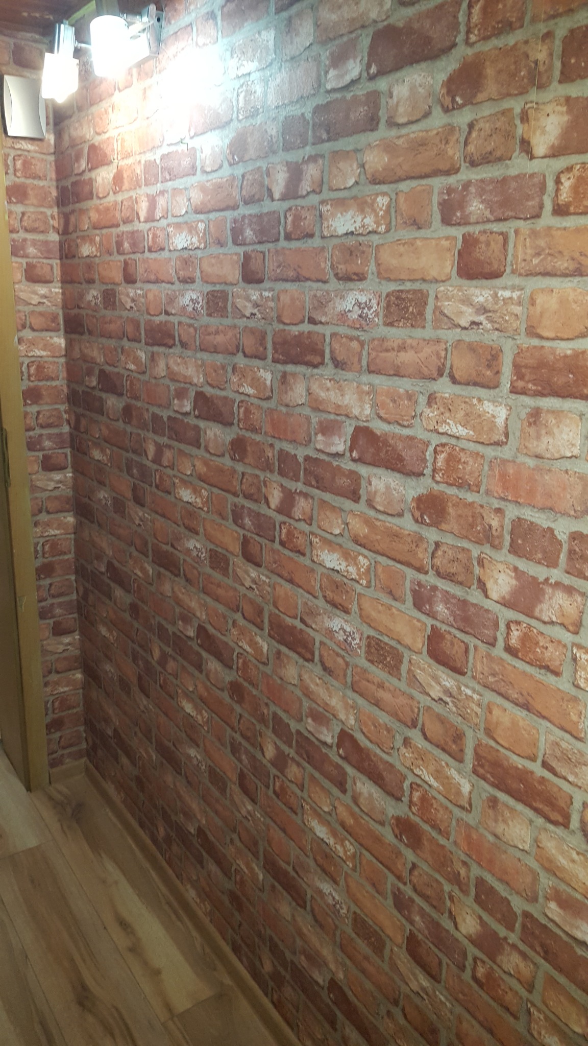 Ściana wykończona tapetą imitującą czerwoną cegłę, oświetlona od góry trzema reflektorami, widoczne drewniane panele podłogowe i fragment ościeżnicy drzwiowej