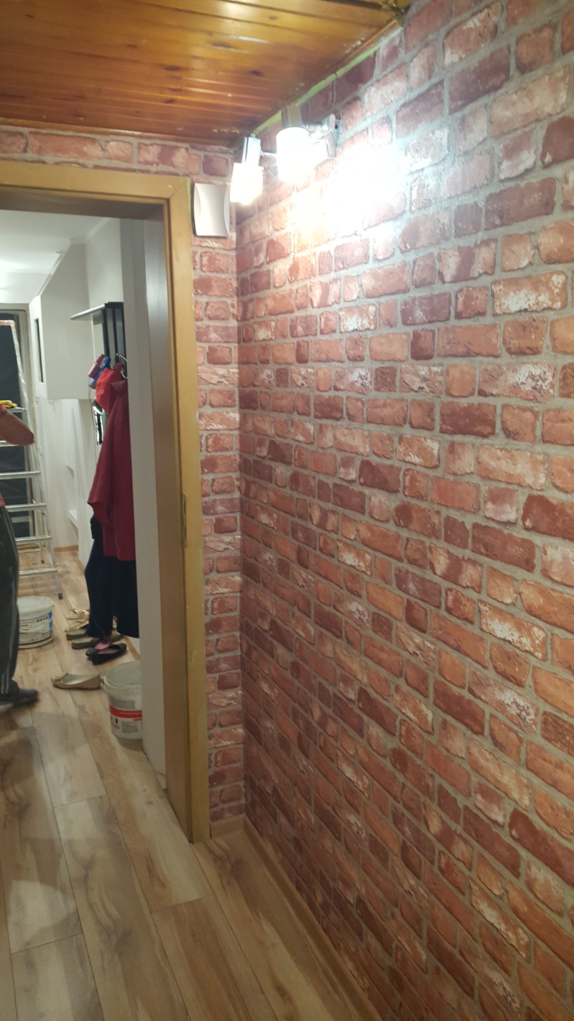 Ściana wykończona tapetą imitującą czerwoną cegłę, oświetlona trzema kinkietami, widoczny fragment drewnianego sufitu i osoby w tle, podłoga wyłożona panelami.