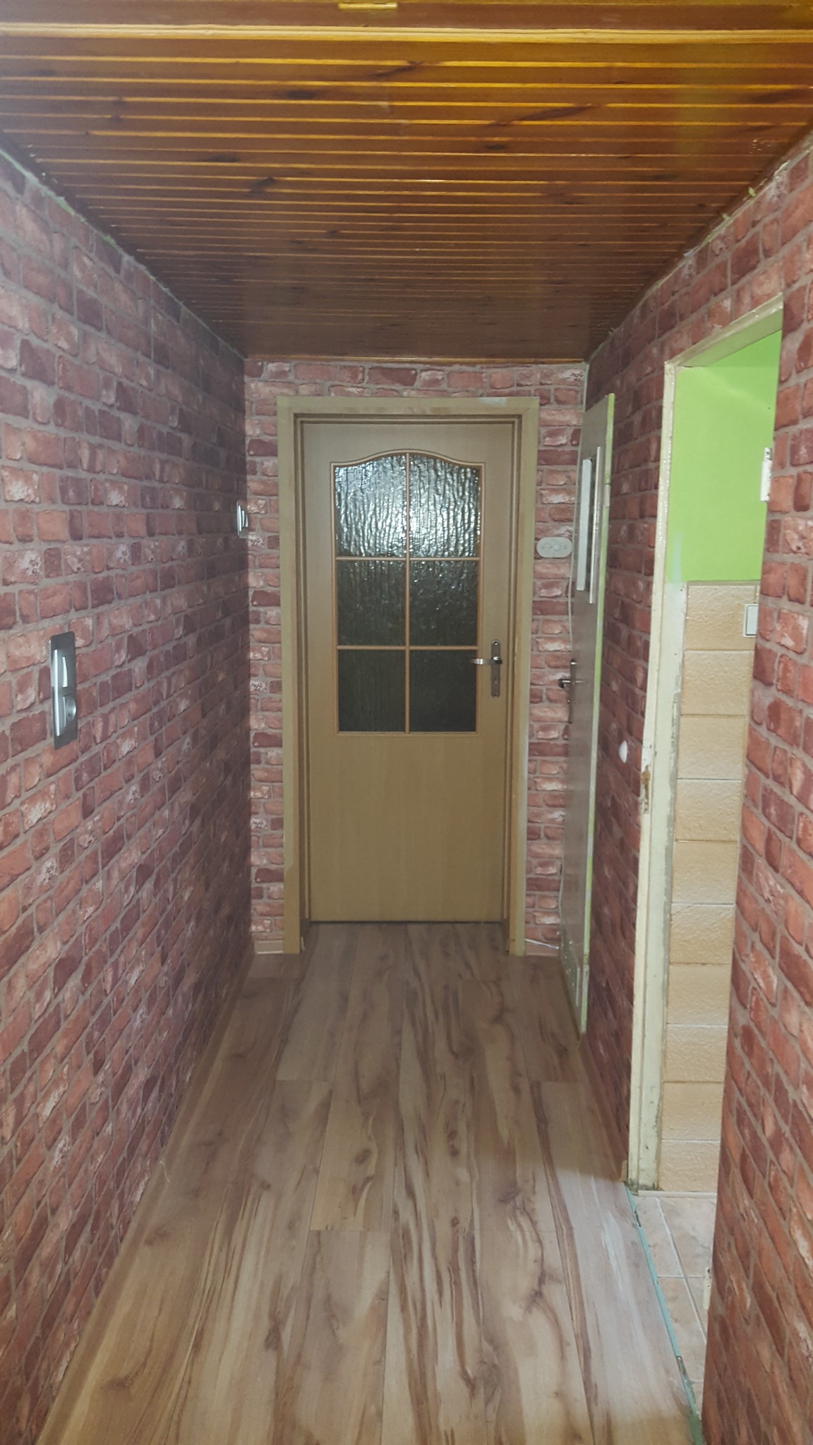 Wąski korytarz z drewnianą podłogą, ścianami wyłożonymi tapetą imitującą czerwoną cegłę i drewnianym sufitem, zakończony beżowymi drzwiami z szybą. Widoczne gniazdko elektryczne na ścianie.