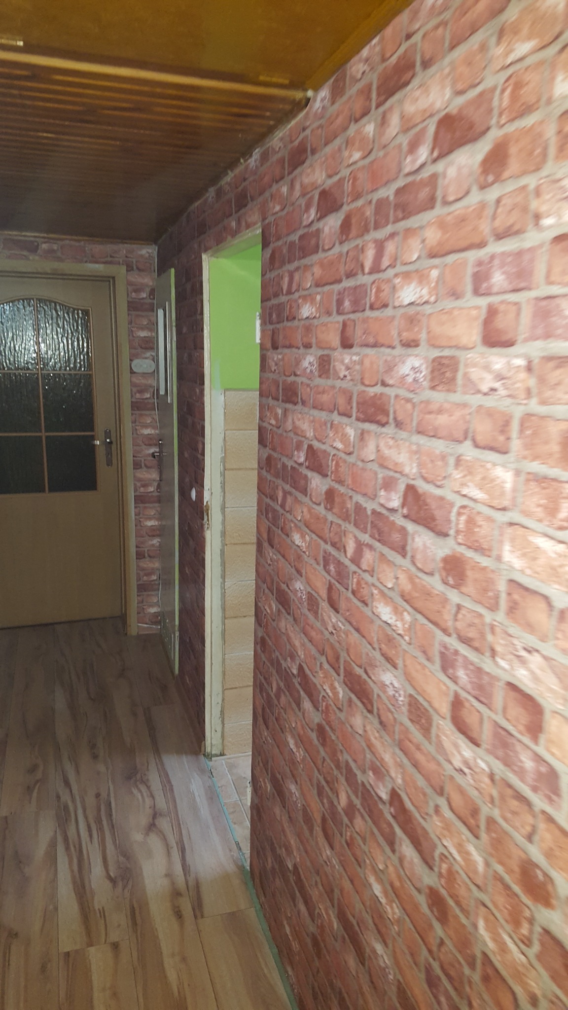 Korytarz po remoncie z drewnianą podłogą, ścianą wykończoną tapetą imitującą czerwoną cegłę i widocznymi drzwiami do pomieszczenia z zieloną ścianą.
