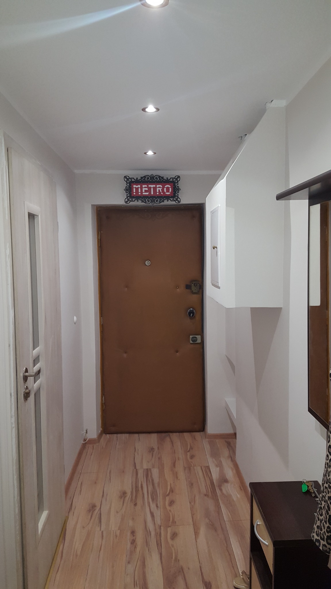 Wnętrze przedpokoju z brązowymi drzwiami wejściowymi, drewnopodobną podłogą i białymi ścianami. Nad drzwiami dekoracyjny szyld 'METRO'.