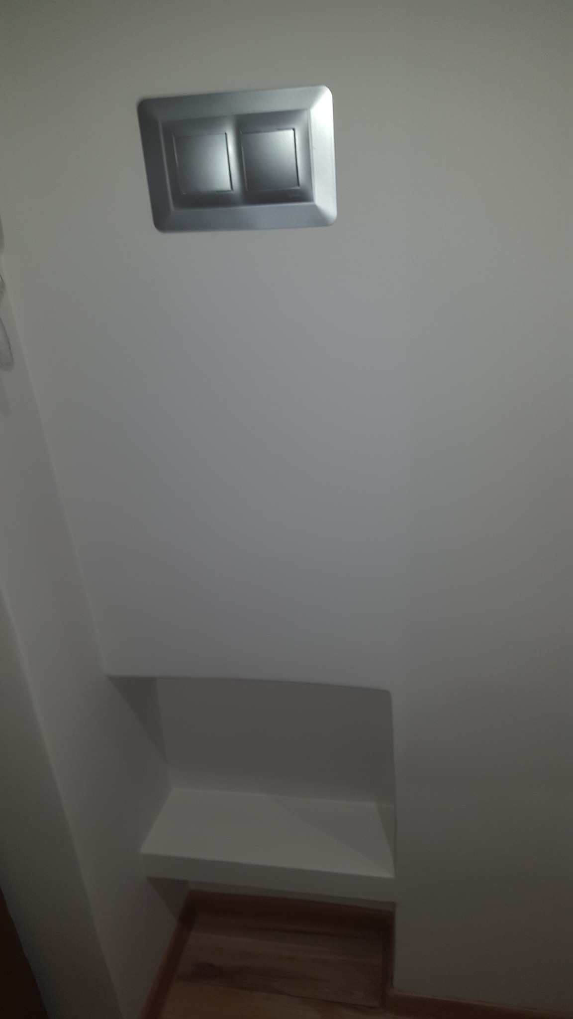 Biała ściana z zamontowanym srebrnym przyciskiem spłuczki i wnęką w dolnej części, widoczny fragment drewnianej podłogi.