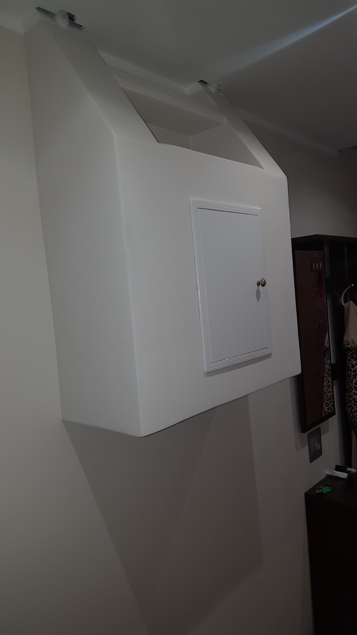 Biała zabudowa sufitowa z małą, kwadratową klapą rewizyjną z ozdobną ramką i złotym uchwytem, widoczna na tle ściany i fragmentu szafy.