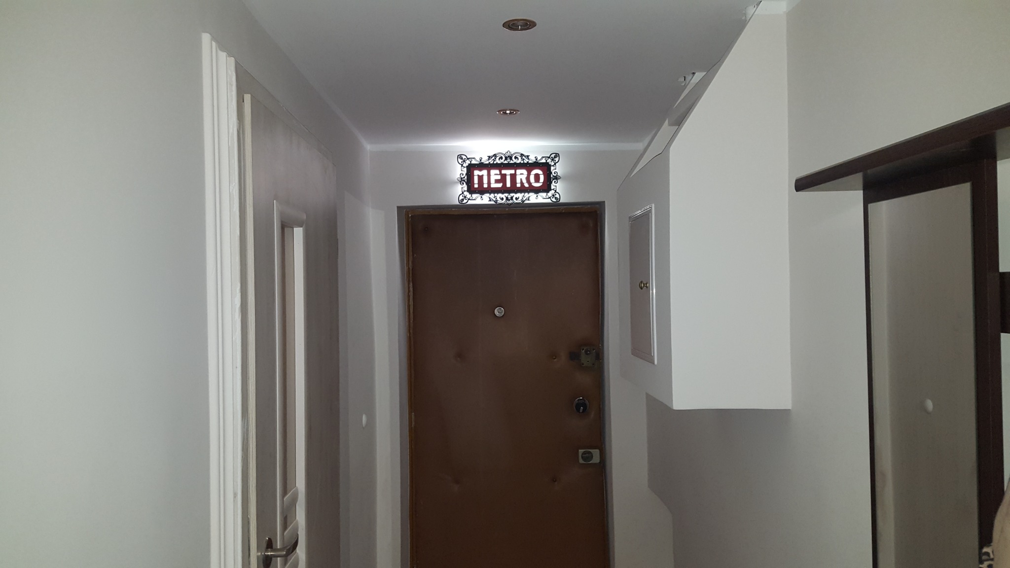 Korytarz z brązowymi drzwiami wejściowymi, nad którymi wisi ozdobny szyld z napisem 'METRO', białe ściany i drzwi po lewej stronie.
