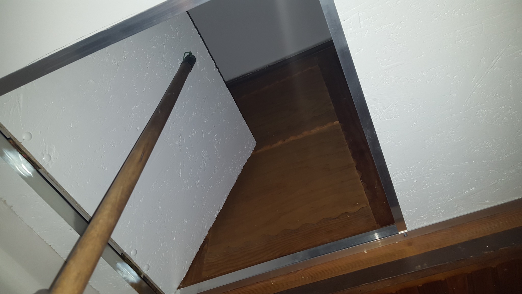 Otwarty właz rewizyjny w suficie z widoczną białą płytą inspekcyjną i drewnianą przestrzenią nad sufitem, oświetloną od góry.