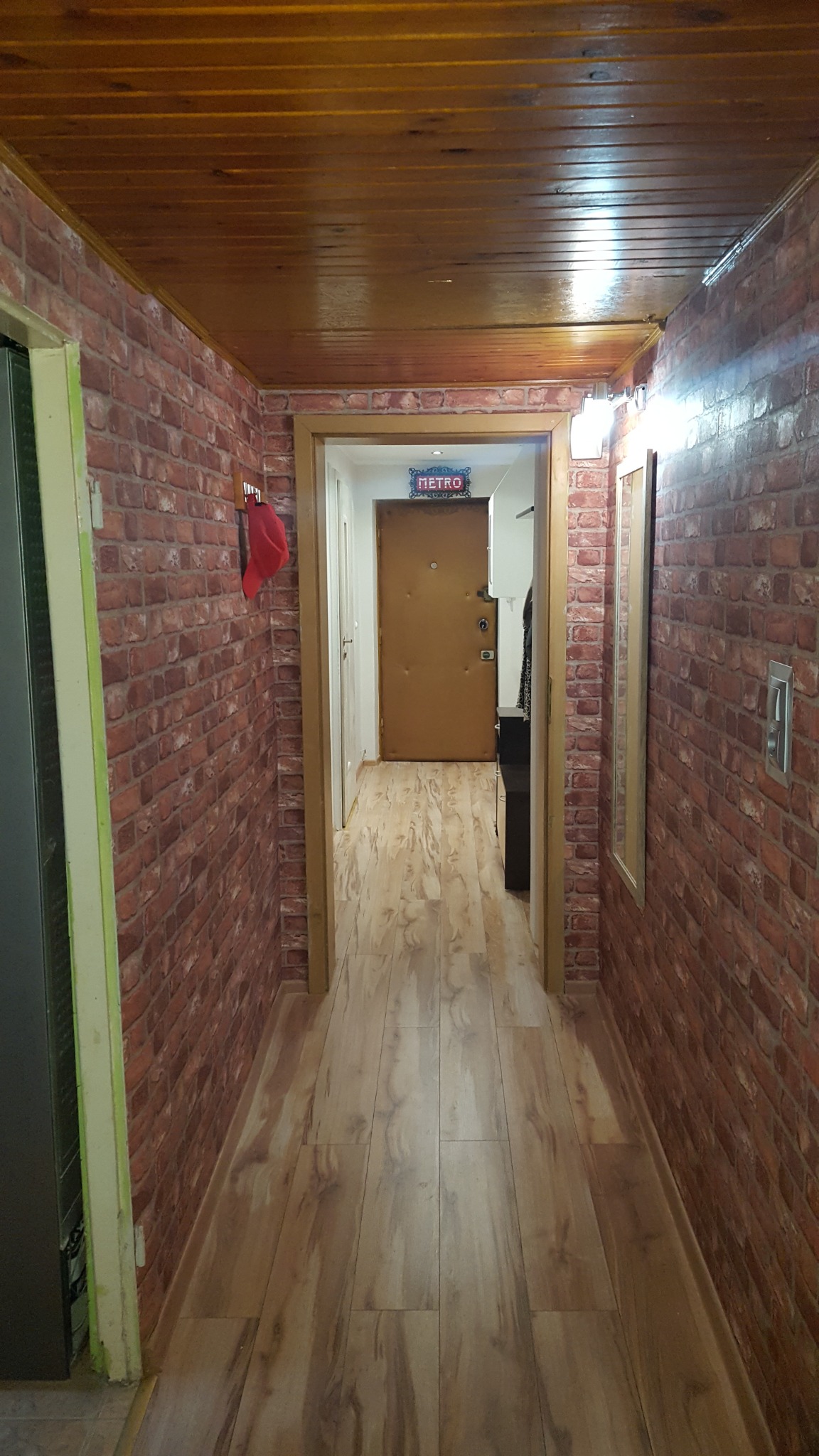 Wąski korytarz z drewnianą podłogą, ścianami z tapetą imitującą czerwoną cegłę i boazerią na suficie; w tle widoczne drzwi wejściowe z podświetlanym napisem 'METRO'.
