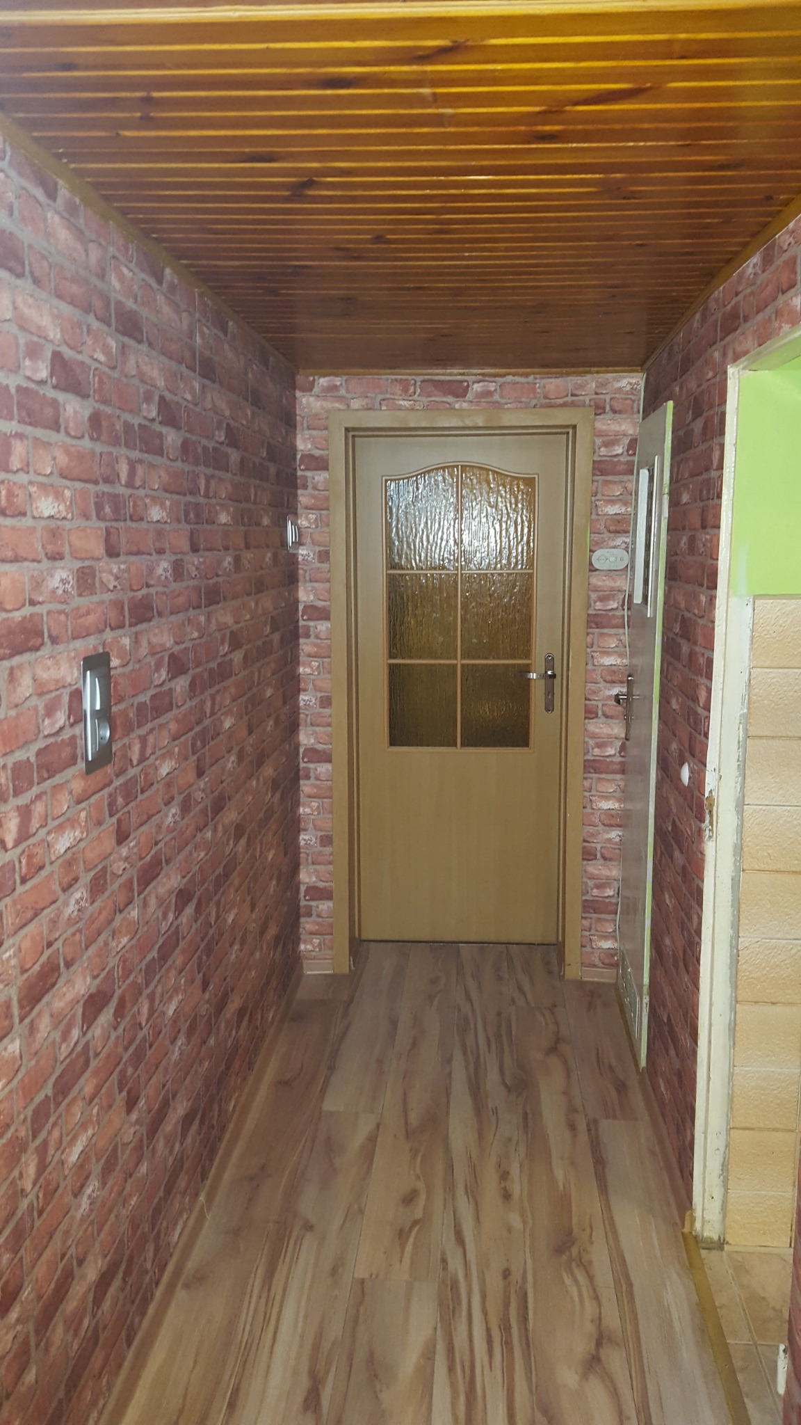 Korytarz z imitacją cegły na ścianach, drewnianą podłogą i sufitem, oraz beżowymi drzwiami z szybą.
