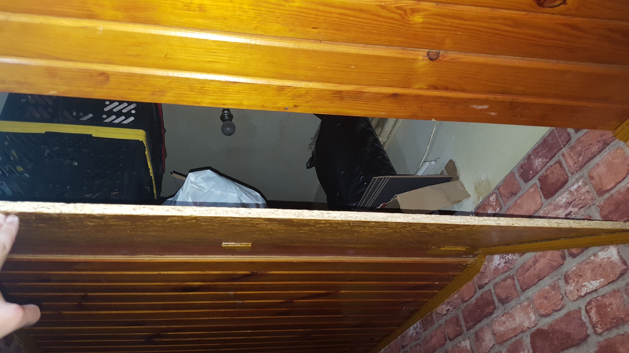 Widok z dołu na drewnianą konstrukcję sufitu z przestrzenią za nią, gdzie widać białą ścianę, ceglaną tapetę, żarówkę i różne przedmioty.