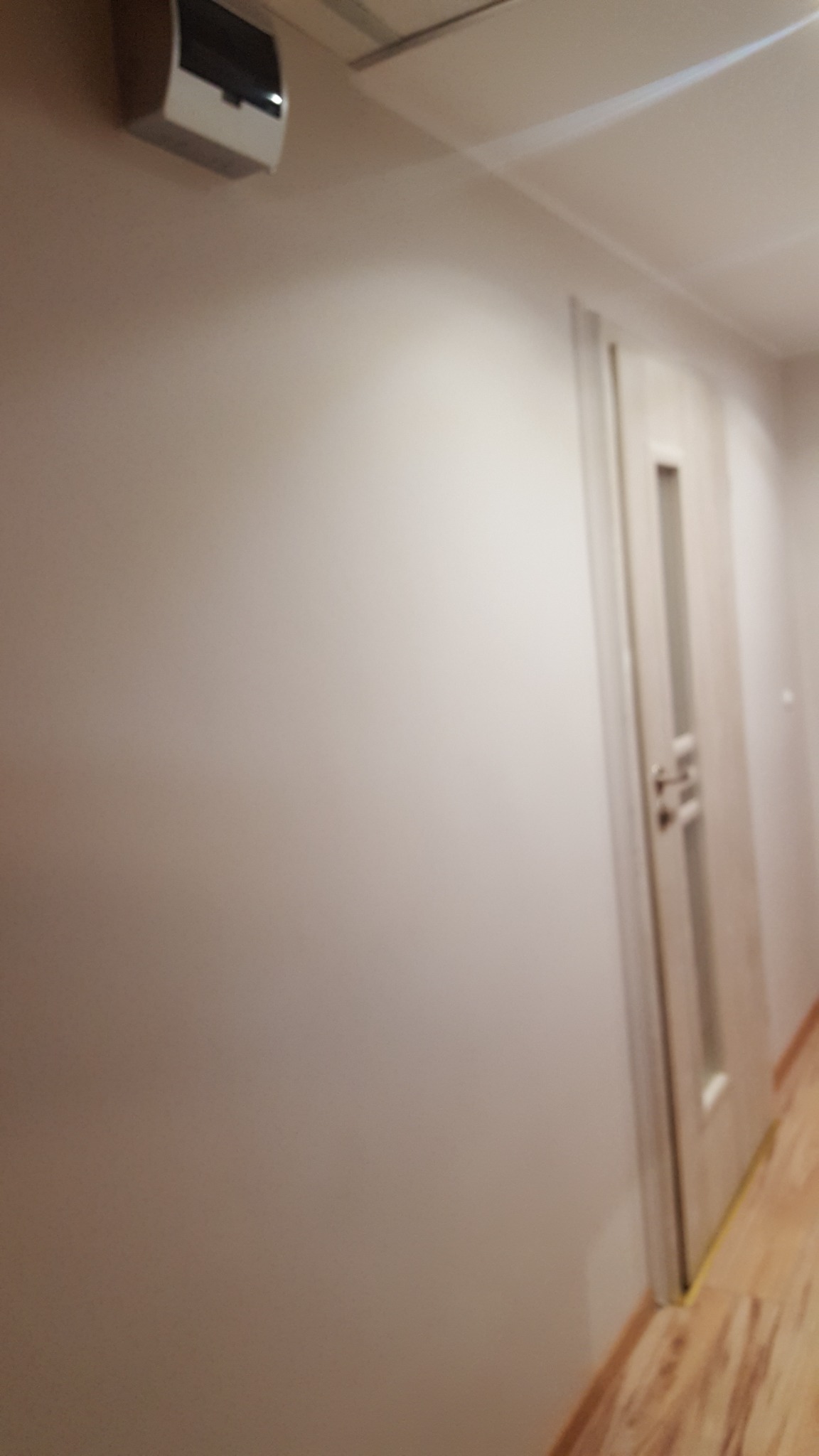 Świeżo pomalowana ściana w jasnym odcieniu w korytarzu, widoczny fragment drzwi z przeszkleniem i listwy przypodłogowe, na górze ściany zamontowana biała skrzynka elektryczna.