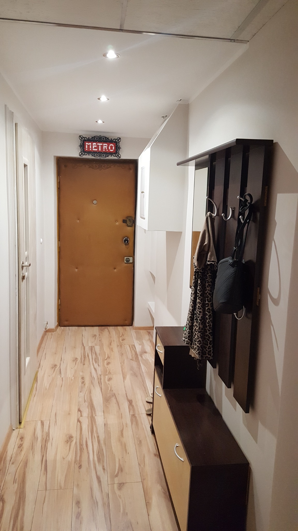 Przedpokój po remoncie: drewnopodobna podłoga, brązowe drzwi wejściowe z dekoracyjnym napisem 'METRO' nad nimi, wieszak z lustrem i szafka na buty.