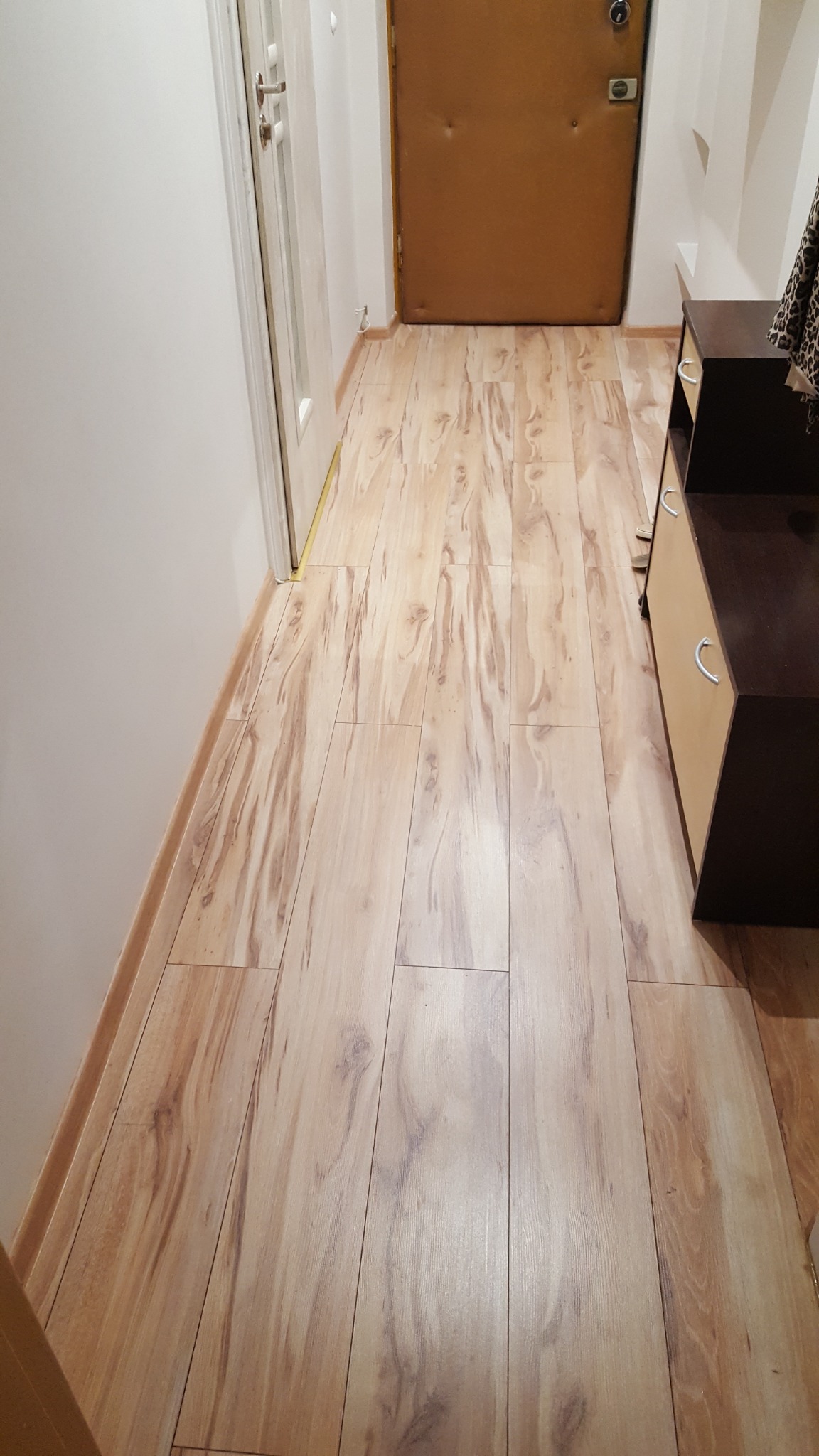Nowa, jasna podłoga laminowana w przedpokoju, imitująca drewno, z widoczną listwą przypodłogową i fragmentem szafki.