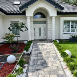 Elegancki dom jednorodzinny z białymi ramami okiennymi, wejściem z kolumnami i szarą kostką brukową prowadzącą do drzwi, otoczony zadbanym ogrodem z kulami dekoracyjnymi i niskimi krzewami.