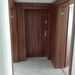 Korytarz z trzema drewnopodobnymi drzwiami; środkowe drzwi z metalową klamką i wizjerem, marmurowa podłoga.