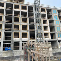 Wznoszenie wielopiętrowego budynku w stanie surowym, z częściowo zamontowanymi oknami zabezpieczonymi folią, widoczny żuraw budowlany i tymczasowe ogrodzenie.