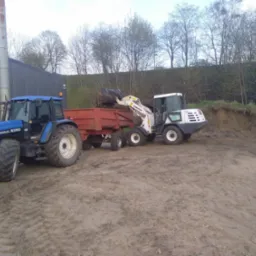 Niebieski traktor New Holland 8360 i biała ładowarka Komatsu WA90 z łyżką, załadowująca ziemię do czerwonej przyczepy na piaszczystym terenie, w tle budynek z blachy falistej i skarpa.