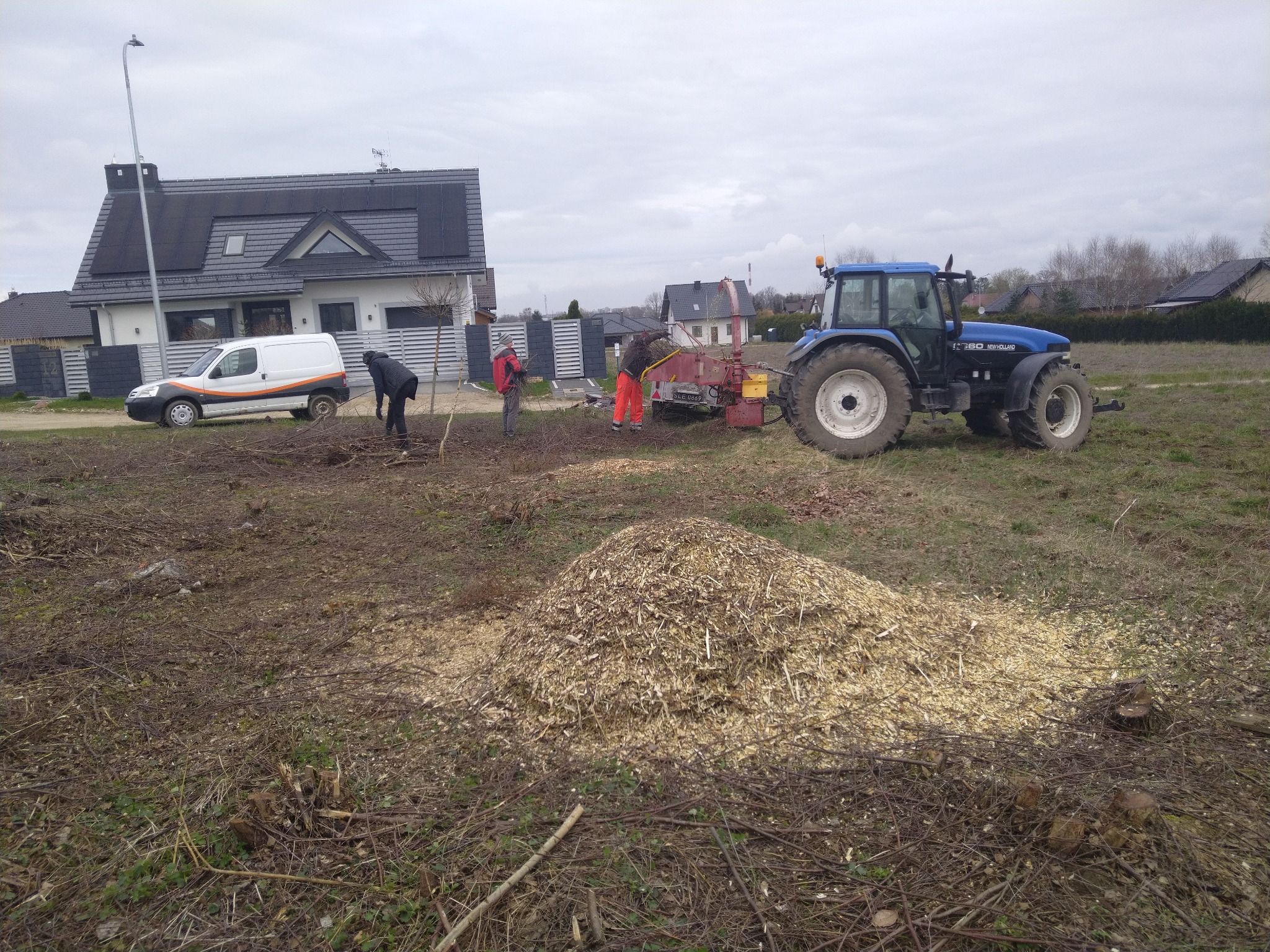 Brygada robotników w trakcie karczowania terenu pod zabudowę, z użyciem traktora i rębaka do gałęzi, na pierwszym planie pryzma zrębków, w tle domy jednorodzinne.