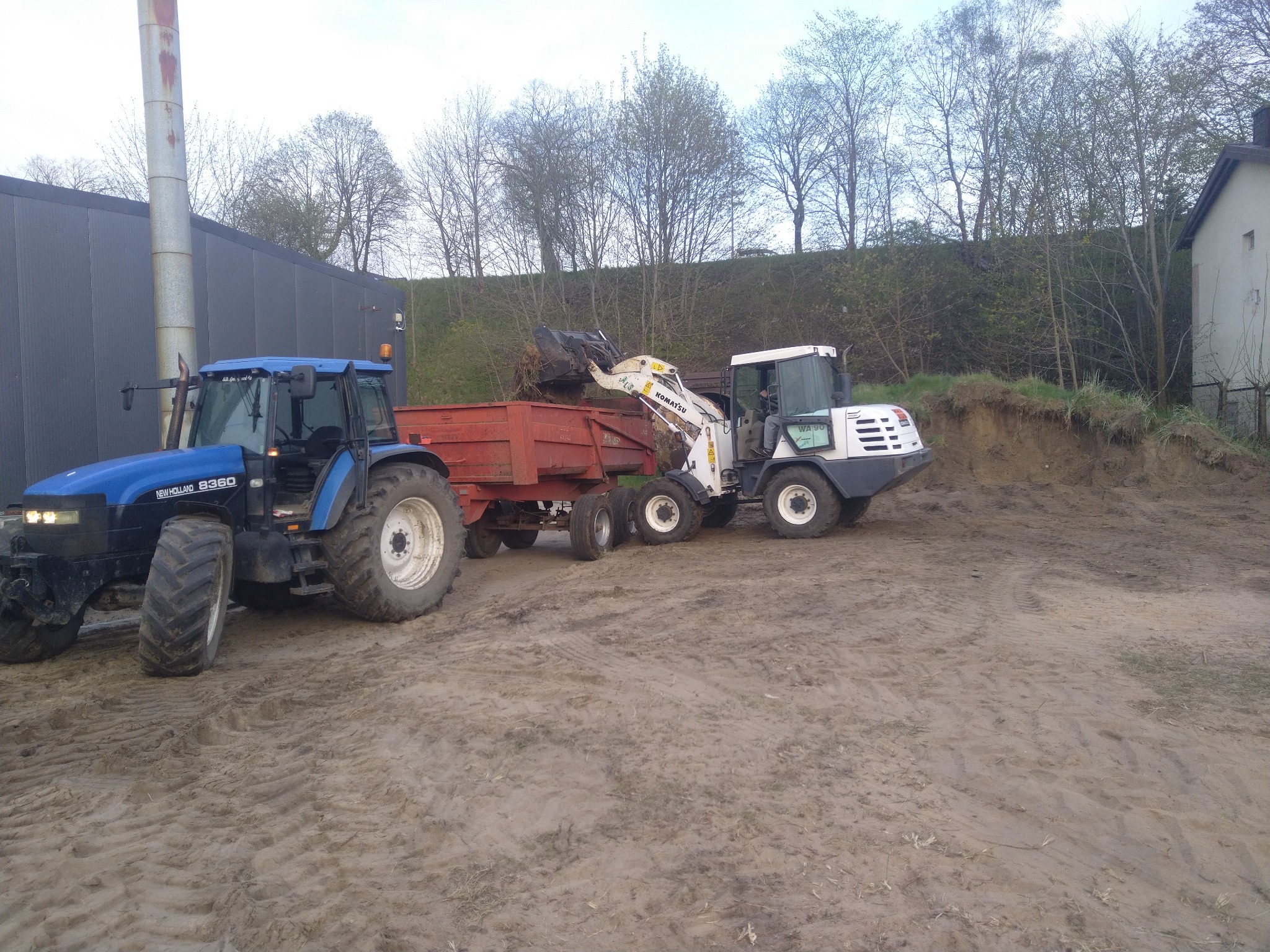 Niebieski traktor New Holland 8360 i biała ładowarka Komatsu WA90 z łyżką, załadowująca ziemię do czerwonej przyczepy na piaszczystym terenie, w tle budynek z blachy falistej i skarpa.