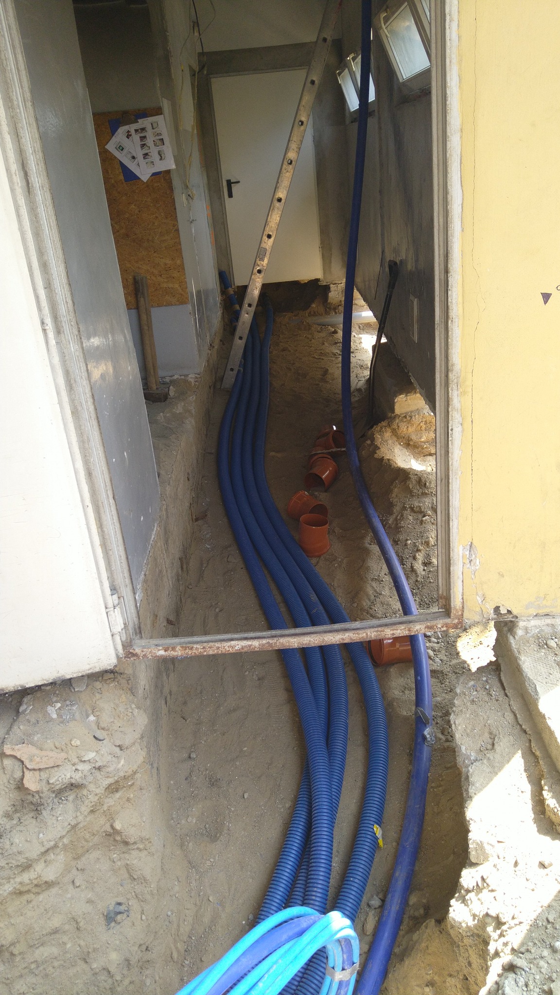 Wykop w gruncie z ułożonymi niebieskimi rurami karbowanymi i pomarańczowymi złączkami, widoczna drabina oparta o ścianę budynku.