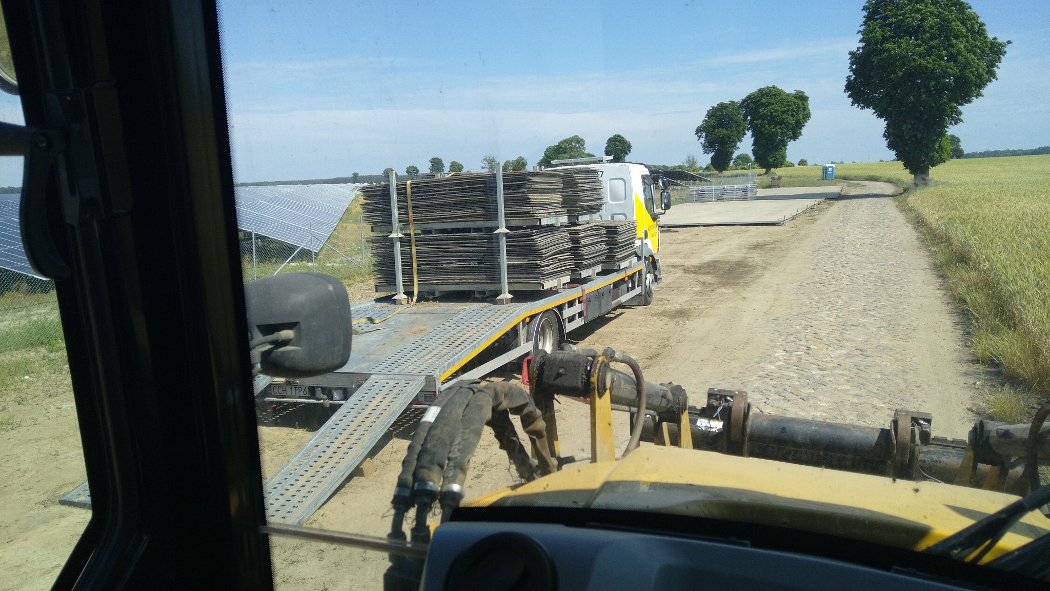 Widok z kabiny maszyny budowlanej na ciężarówkę z przyczepą transportującą płyty utwardzające drogę, w tle panele słoneczne i polna droga.