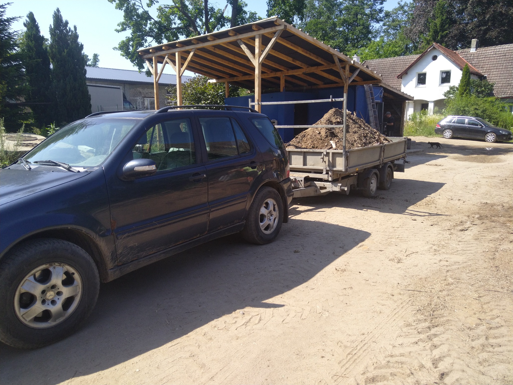 Ciemny SUV Mercedes ciągnie przyczepę wypełnioną ziemią na piaszczystym podwórku, w tle drewniana wiata, dom i czarny kot.
