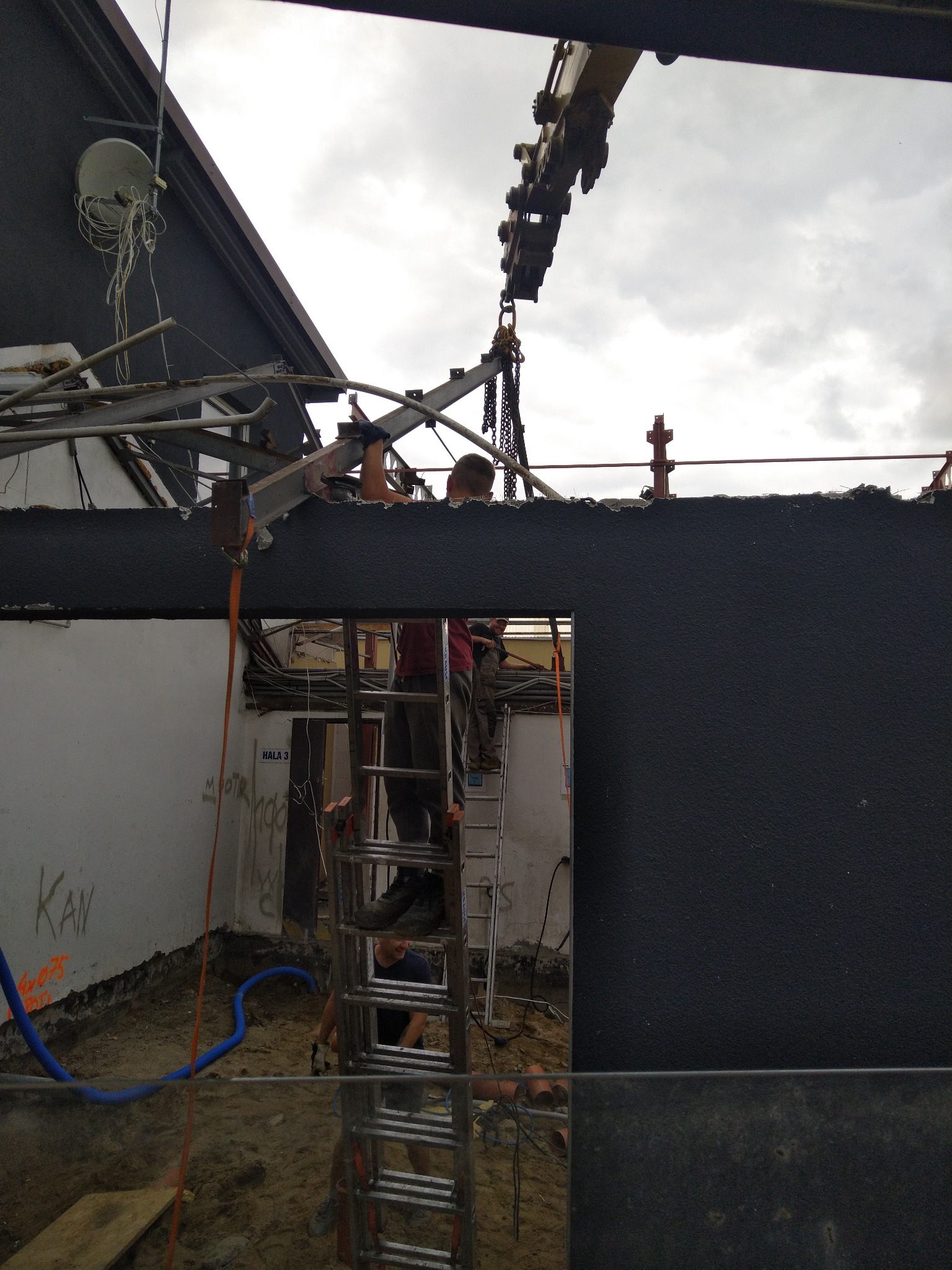 Demontaż konstrukcji budynku z użyciem dźwigu, pracownicy na drabinie, widoczne elementy konstrukcyjne, ściana z napisem, niebieskie rury na ziemi.