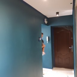 M BUD - Świeżo pomalowany korytarz w odcieniach turkusu, widoczna brązowa, zabezpieczona folią drzwi wejściowe, biała podłoga i pracownik w tle.