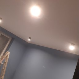 M BUD - Świeżo pomalowane na szaro ściany z zamontowanymi reflektorami sufitowymi, widoczna drabina malarska w rogu pokoju.