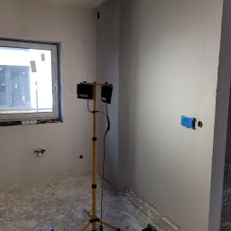 M BUD - Pokój w trakcie remontu: świeżo pomalowane ściany, zaklejone gniazdko, lampa budowlana na statywie, resztki farby na podłodze i listwa przypodłogowa leżąca obok schodów.