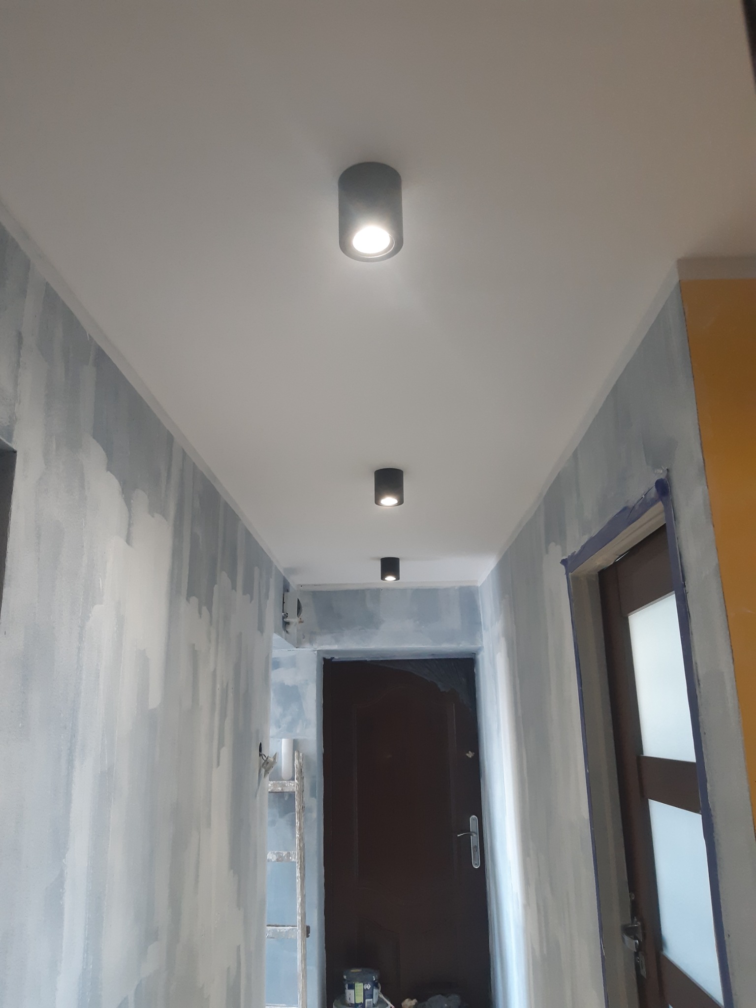 Korytarz w trakcie malowania z widocznymi śladami farby na ścianach, zamontowanymi trzema okrągłymi lampami sufitowymi w kolorze grafitowym oraz drabina malarska oparta o ścianę.