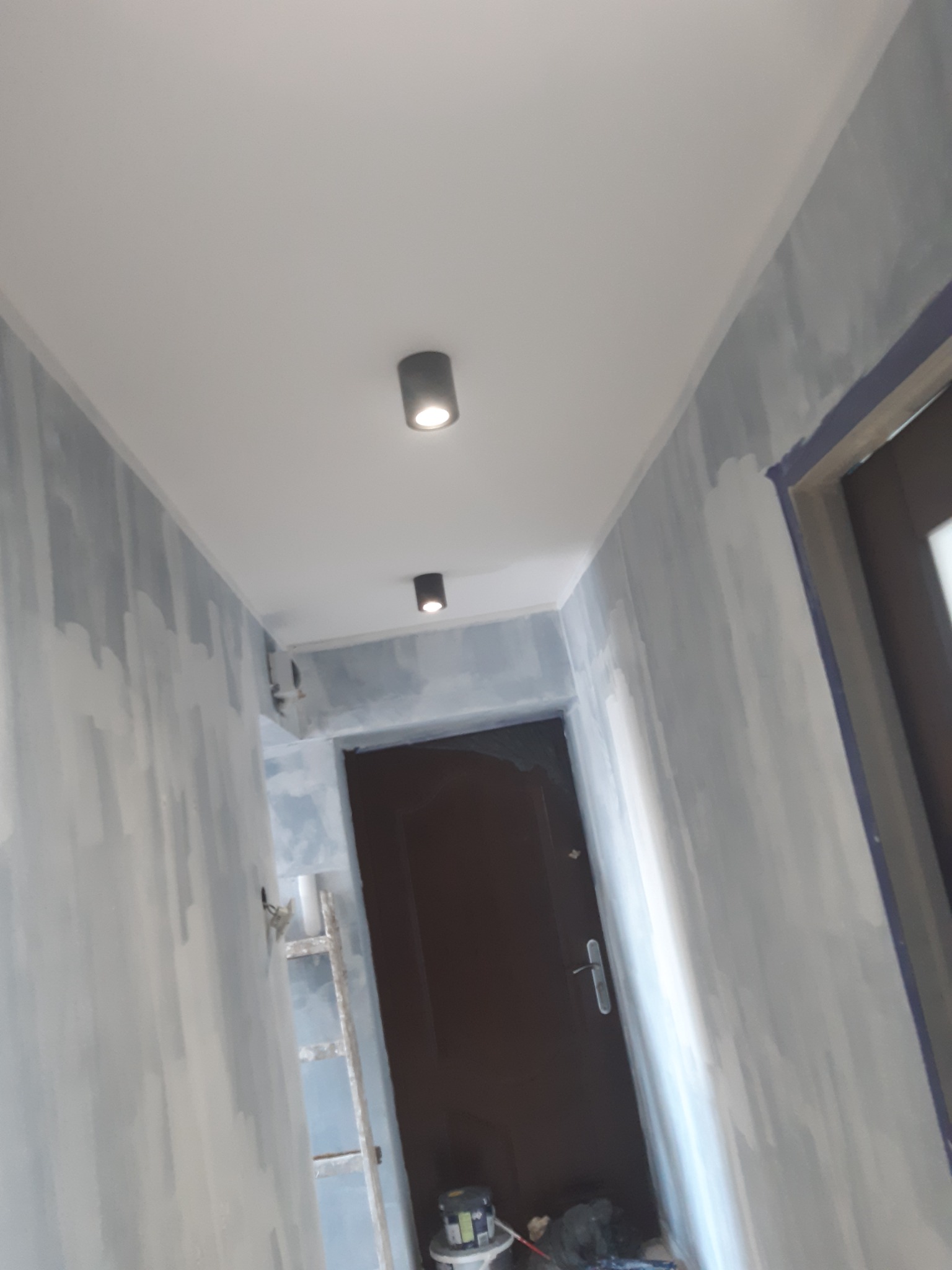 Korytarz w trakcie malowania: ściany pokryte pierwszą warstwą jasnoniebieskiej farby, widoczna drabina oparta o ścianę, dwie czarne lampy sufitowe punktowe, drzwi wejściowe i wiadro z farbą w rogu.