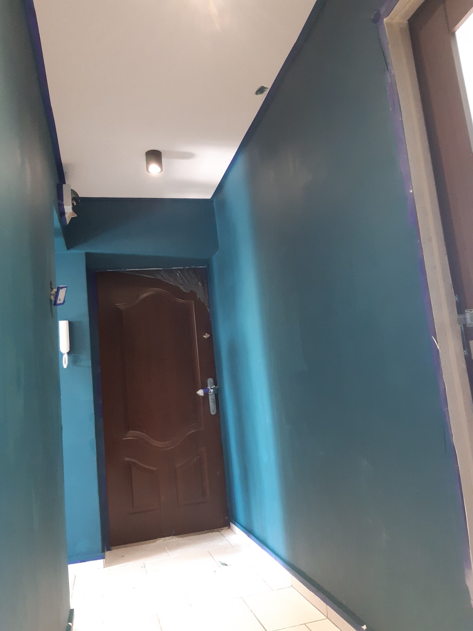 Korytarz po malowaniu na kolor morski, widoczna brązowa okleina drzwi zabezpieczona folią malarską, biały sufit z punktowym oświetleniem, ślady taśmy malarskiej wzdłuż krawędzi.