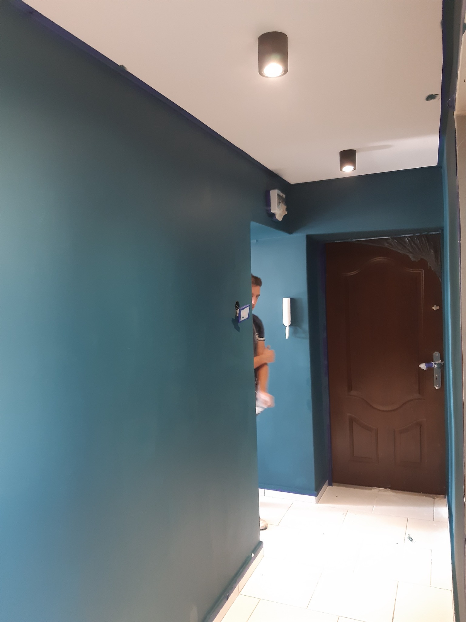Świeżo pomalowany korytarz w odcieniach turkusu, widoczna brązowa, zabezpieczona folią drzwi wejściowe, biała podłoga i pracownik w tle.