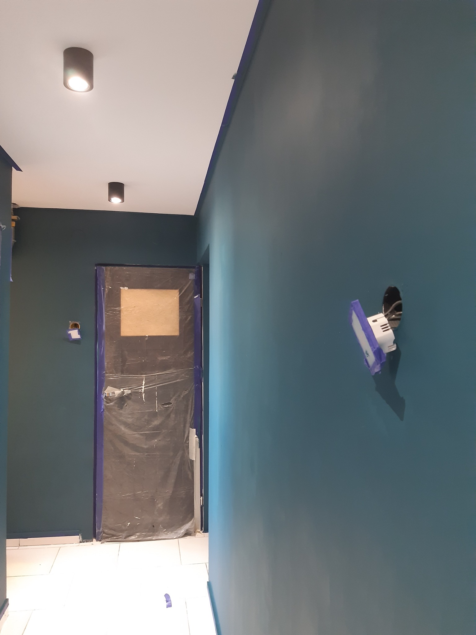 Świeżo pomalowane, ciemnozielone ściany w korytarzu, z zabezpieczoną folią i taśmą malarską ościeżnicą drzwi oraz puszką elektryczną przygotowaną do montażu osprzętu.