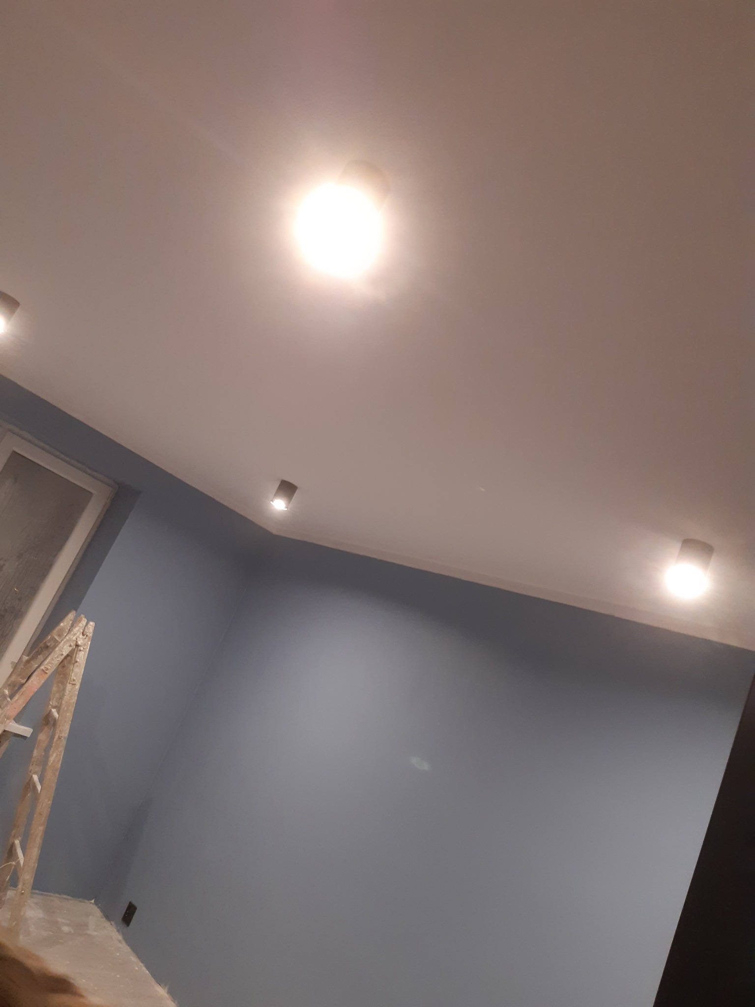 Świeżo pomalowane na szaro ściany z zamontowanymi reflektorami sufitowymi, widoczna drabina malarska w rogu pokoju.